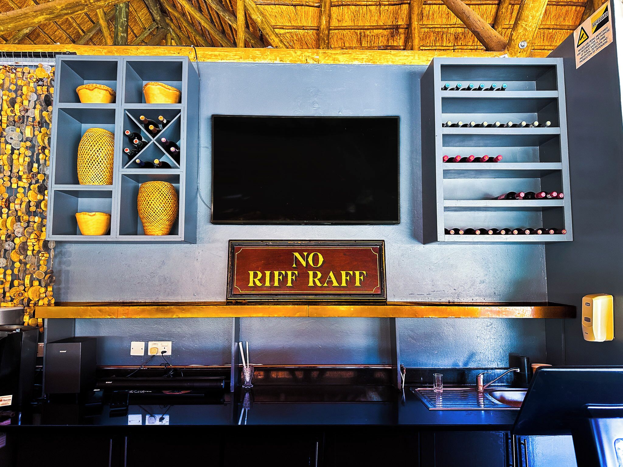 Zona de bar con un cartel que dice 'NO RIF RAFF', estantes con botellas y una pantalla grande.