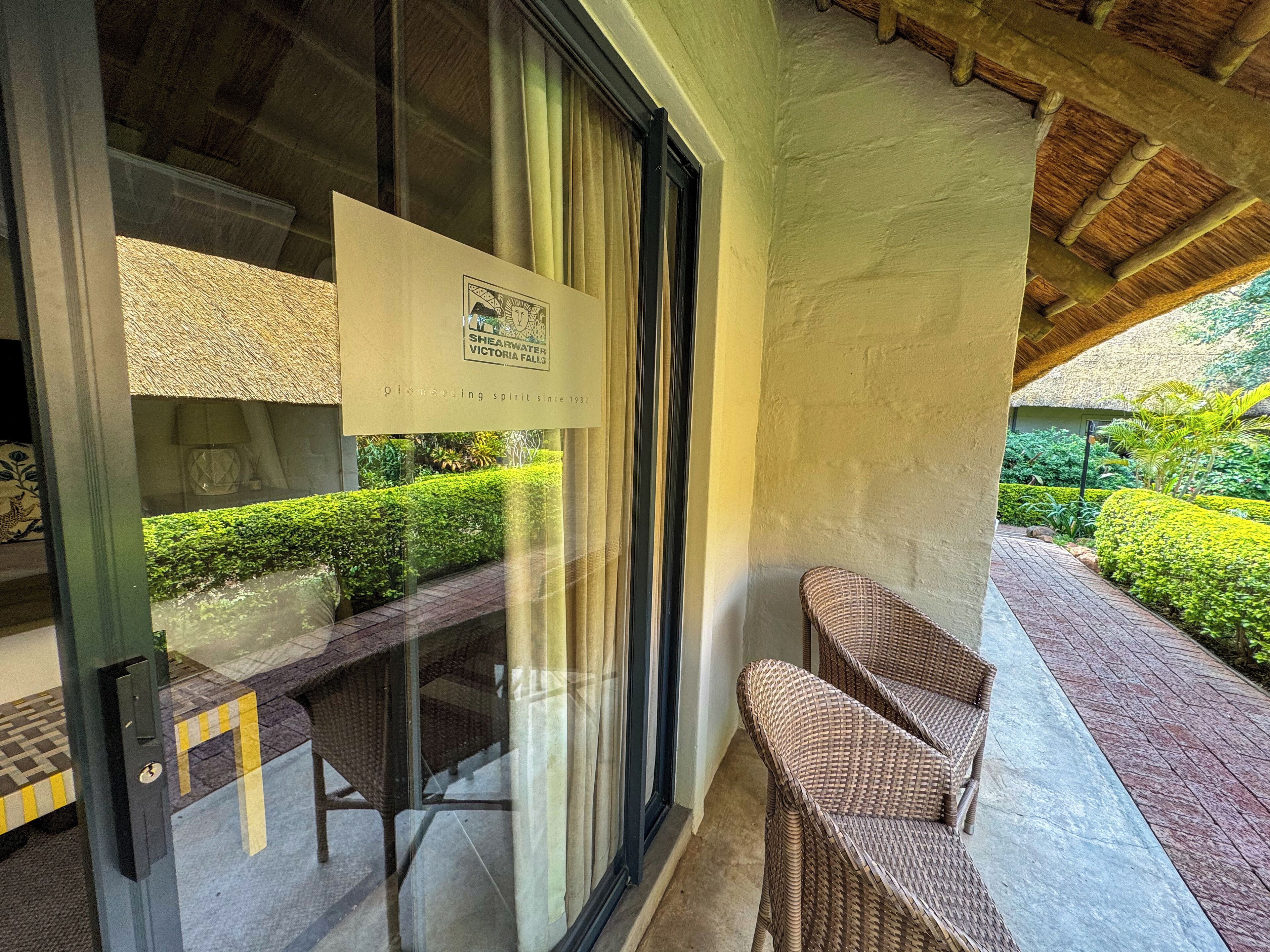 A glass door with a sign for Shearwater Victoria Falls, wicker furniture, and a well-maintained garden outside.