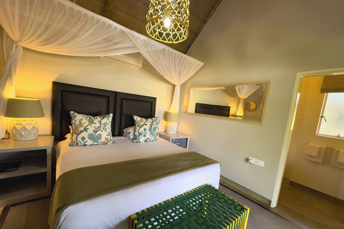 Quarto de hotel com cama, abajures, mesa de cabeceira, espelho e janela.