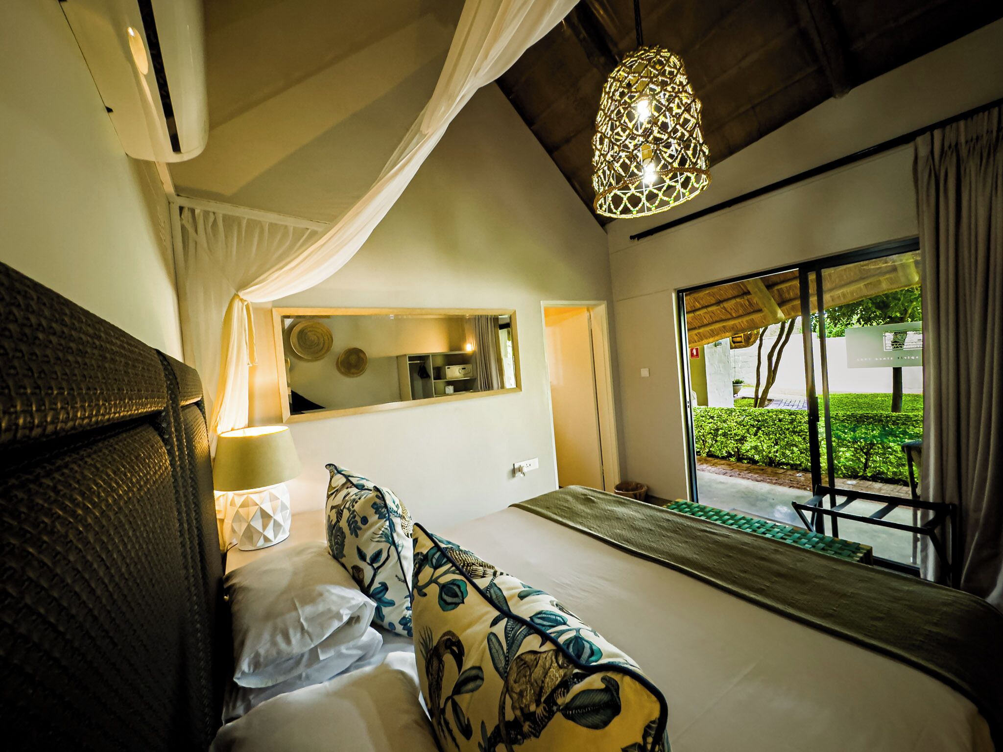 A bedroom with a bed, a mirror, a hanging light fixture, and a view of the outdoors.