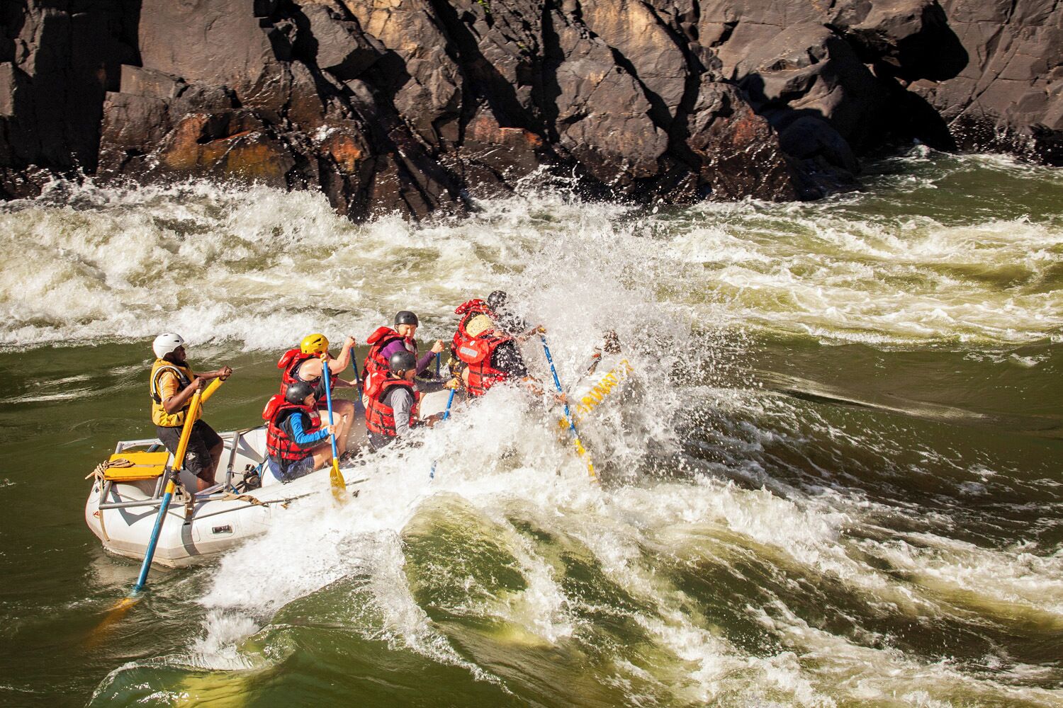 A group of people in a white raft navigating through a river with rapids, surrounded by rocky cliffs.