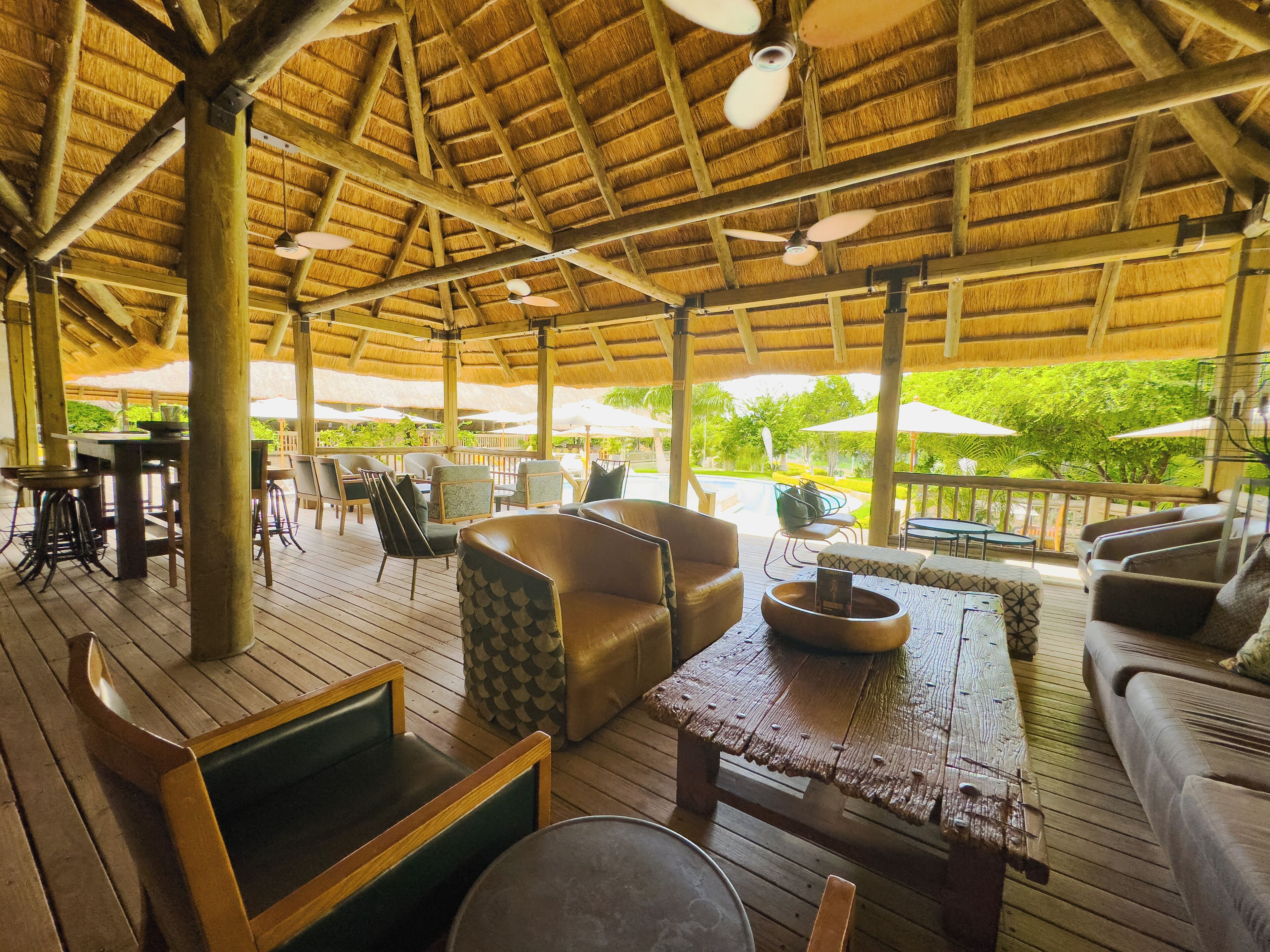An outdoor lounge area with wooden flooring, seating arrangements including sofas and chairs, a wooden table, and a thatched roof structure.