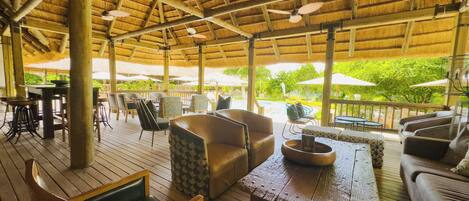 An outdoor lounge area with wooden flooring, seating arrangements including sofas and chairs, a wooden table, and a thatched roof structure.