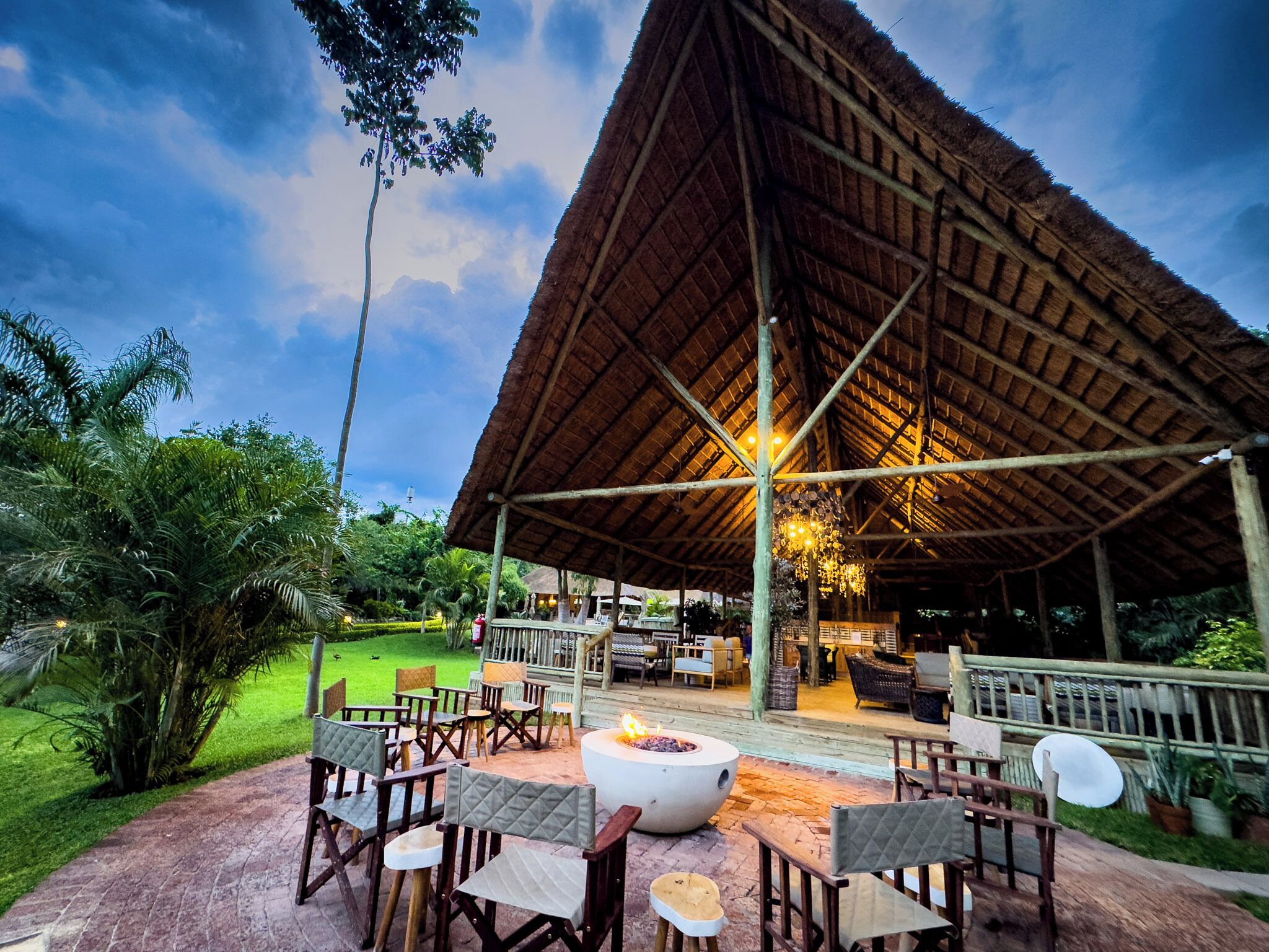 An outdoor seating area with a fire pit, surrounded by wooden chairs and tables, under a thatched roof structure.