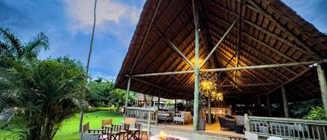 An outdoor seating area with a fire pit, surrounded by wooden chairs and tables, under a thatched roof structure.