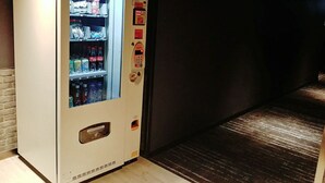 Máquina expendedora con diversas bebidas y aperitivos, ubicada en un pasillo con piso oscuro y pared de ladrillo.