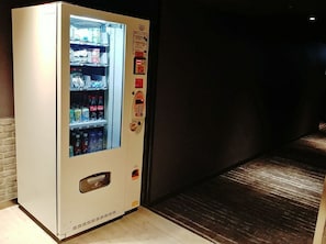 A vending machine with a variety of beverages and snacks, located in a hallway with a dark floor and a brick wall.