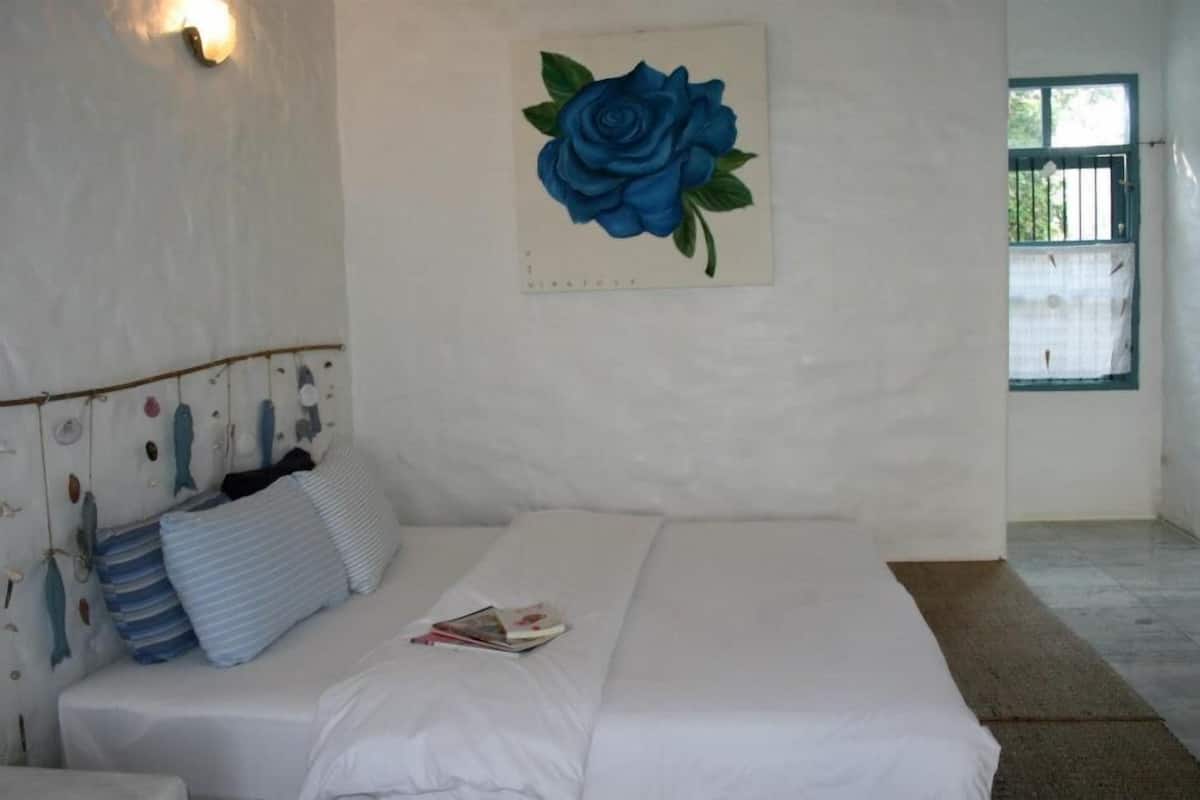 A bedroom with a bed, a wall-mounted painting of a blue rose, a window, and a wall with a light fixture.
