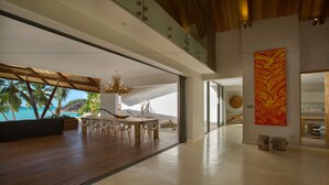 Un interior moderno con una zona de comedor de madera, ventanas amplias con vista a la playa y un cuadro abstracto llamativo.