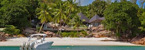 A beachfront property with a boat docked in the water, surrounded by lush greenery and palm trees. - Villa Deckenia (Praslin Island)