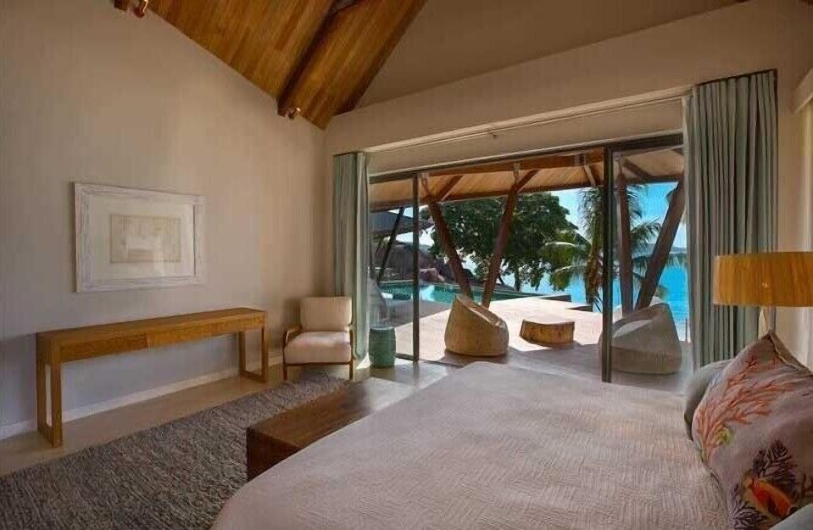 Ein Schlafzimmer mit einem großen Bett, einer Bank, einem Sessel und Blick auf einen Pool und Palmen.