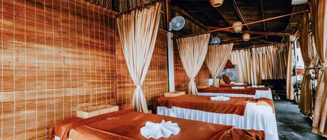 A row of massage tables with white linens and brown towels, set against a bamboo wall with curtains.