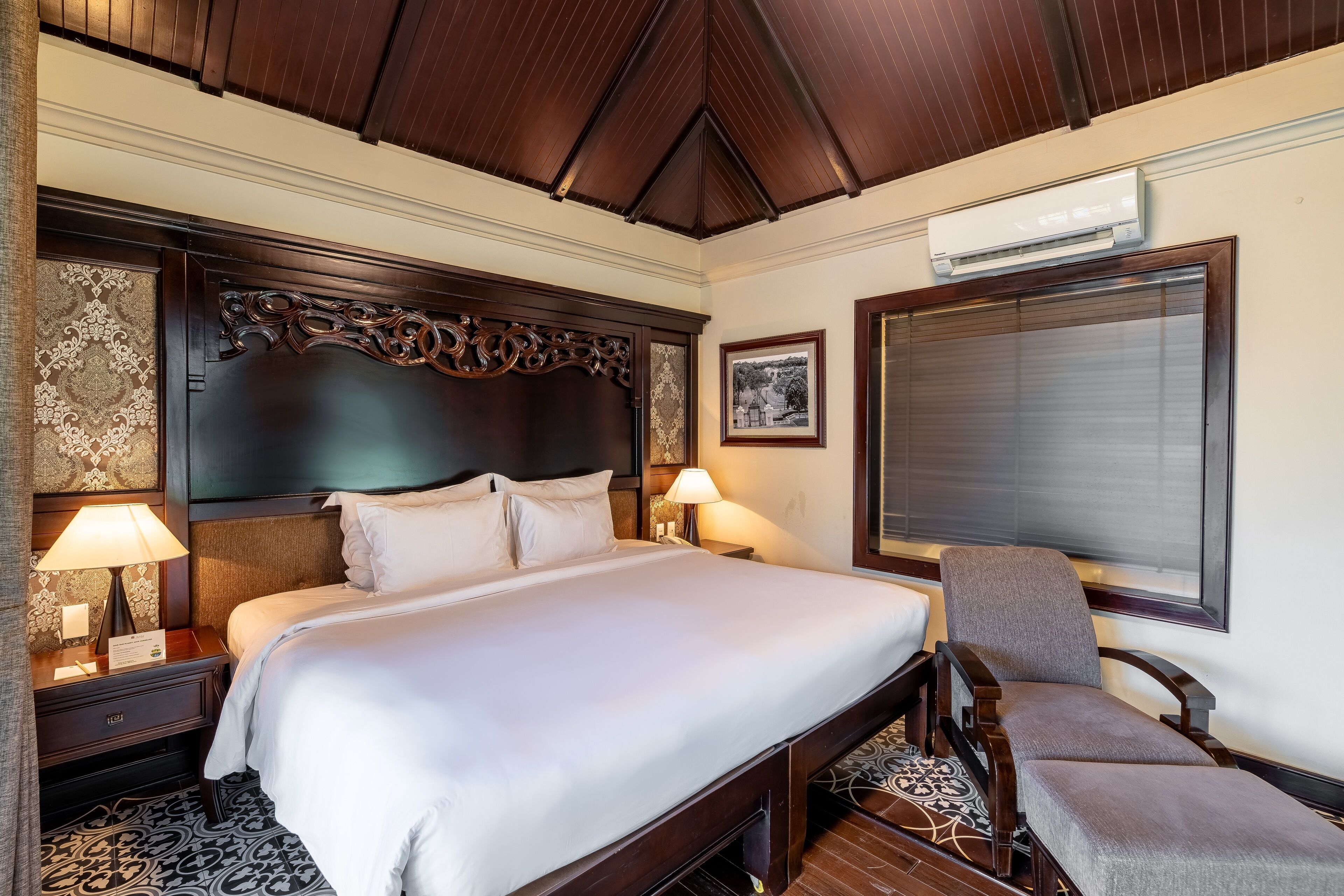 Habitación de hotel con una cama grande, dos mesitas de noche con lámparas, una silla, un ventanal con persianas y un equipo de aire acondicionado.