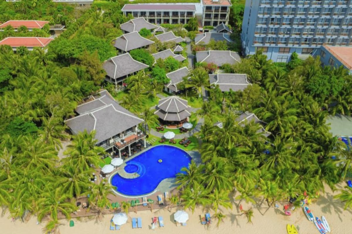 Ein Resort mit mehreren bungalowsartigen Gebäuden, einem zentralen Schwimmbad und einem Strandbereich mit Liegestühlen.