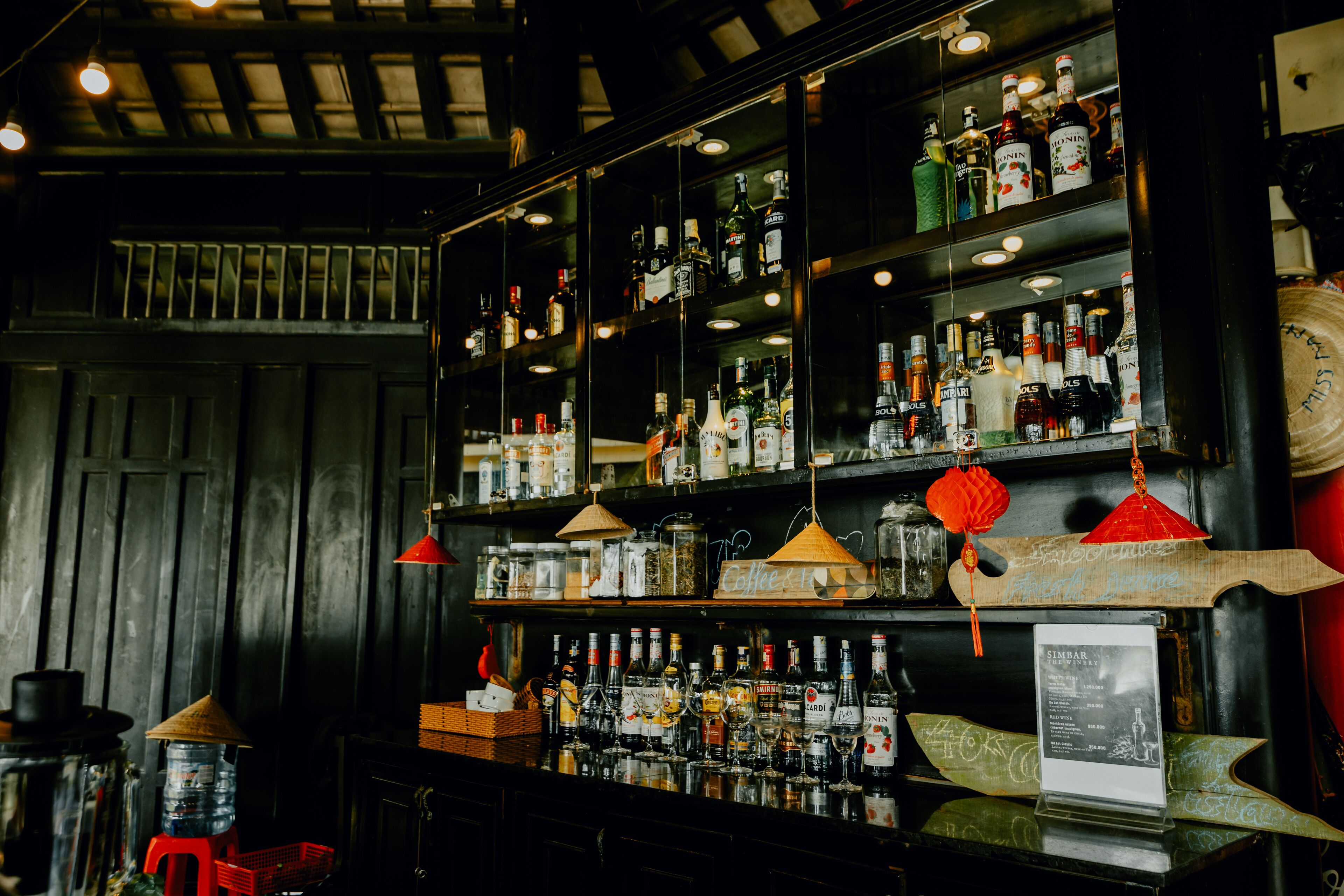 Un bar avec des étagères remplies de bouteilles et de verres de toutes sortes, un présentoir à menus et des éléments de décoration traditionnels, comme une enseigne en bois et des lanternes rouges.