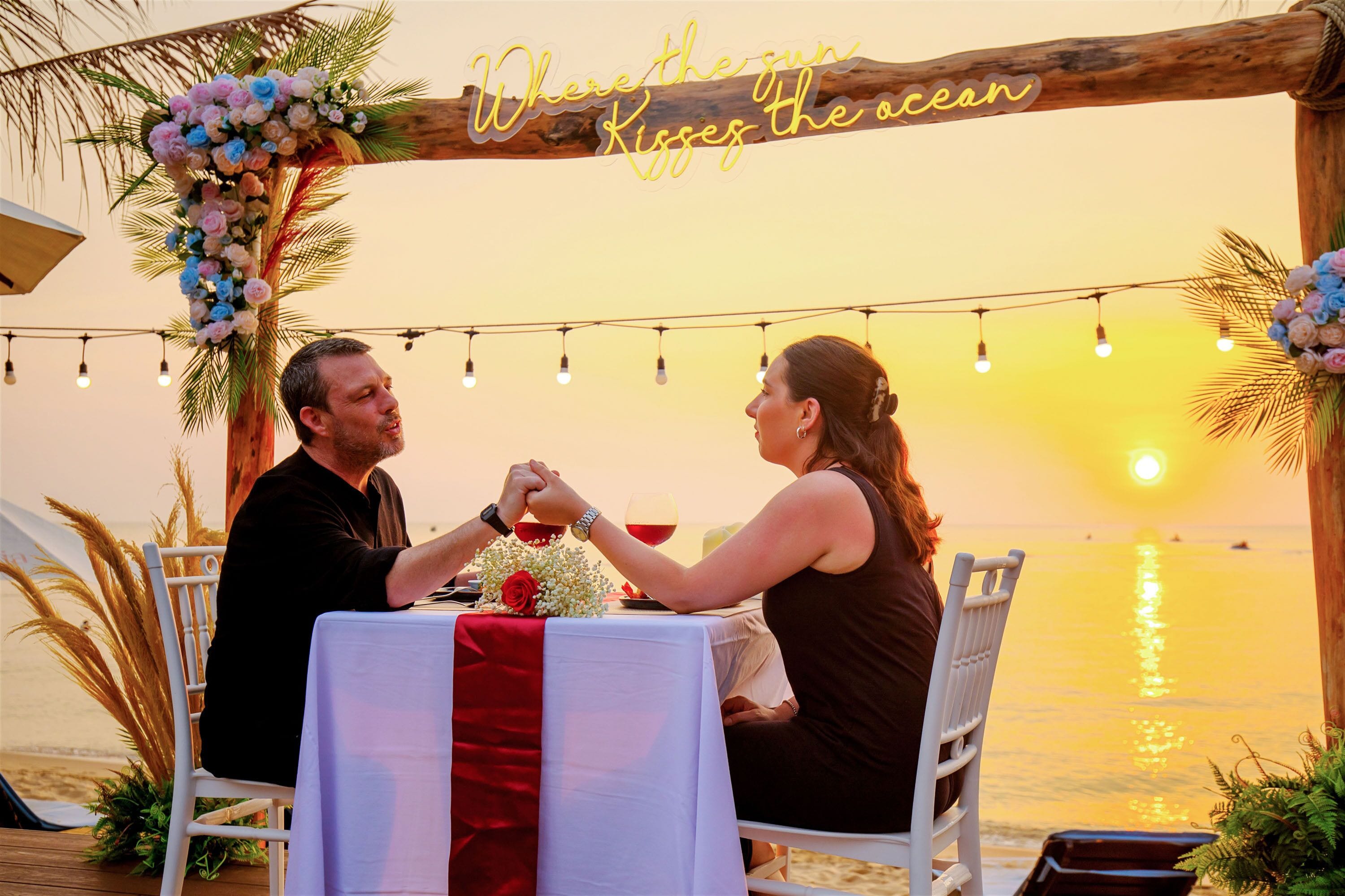 A couple dining at a table with a view of the sunset over the ocean, decorated with flowers and string lights.