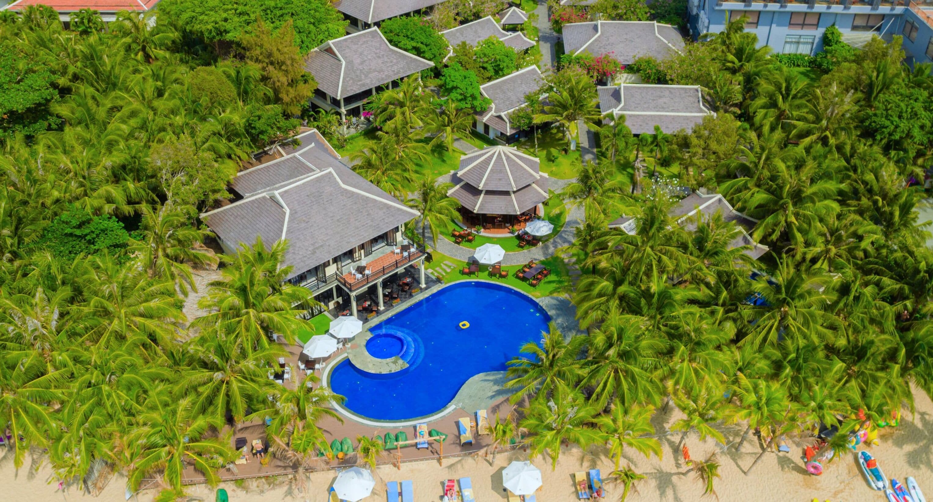 Eine Vogelperspektive zeigt ein Resort mit mehreren bungalowartigen Gebäuden, einem zentralen Swimmingpool und umliegenden Palmen.