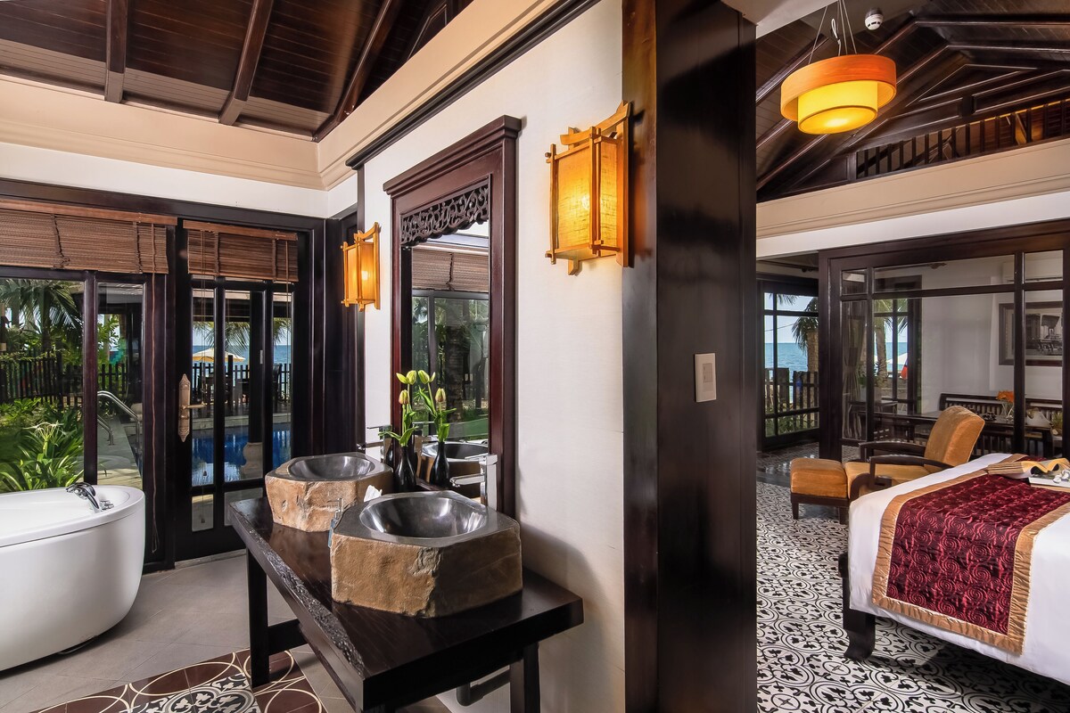 A hotel room with a large bathtub, a wooden vanity with two sinks, a bed with a red blanket, and a balcony with a view of the sea.
