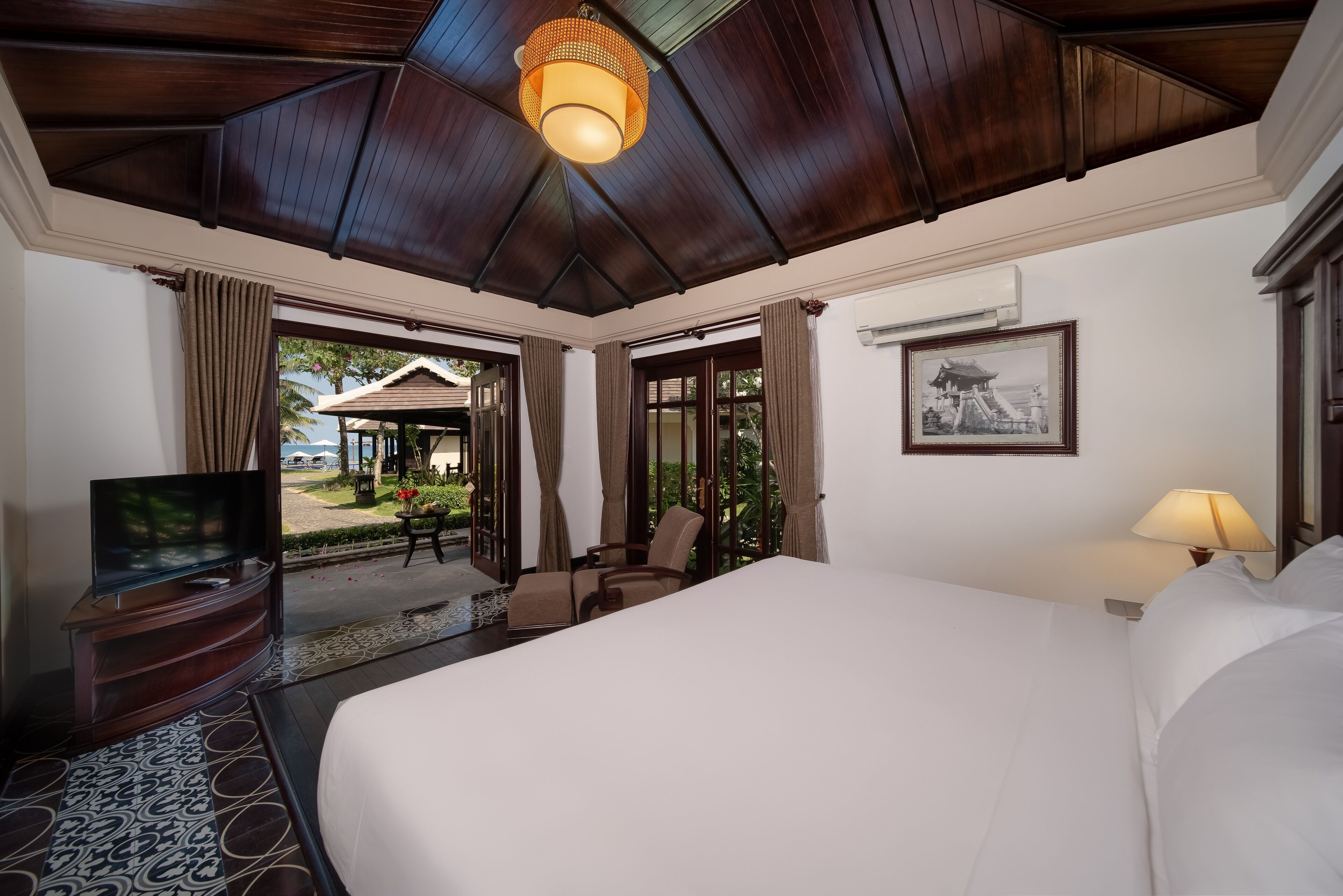 Een hotelkamer met een groot bed, een televisie op een houten standaard, een lamp en uitzicht op een buitenruimte met een gazebo.