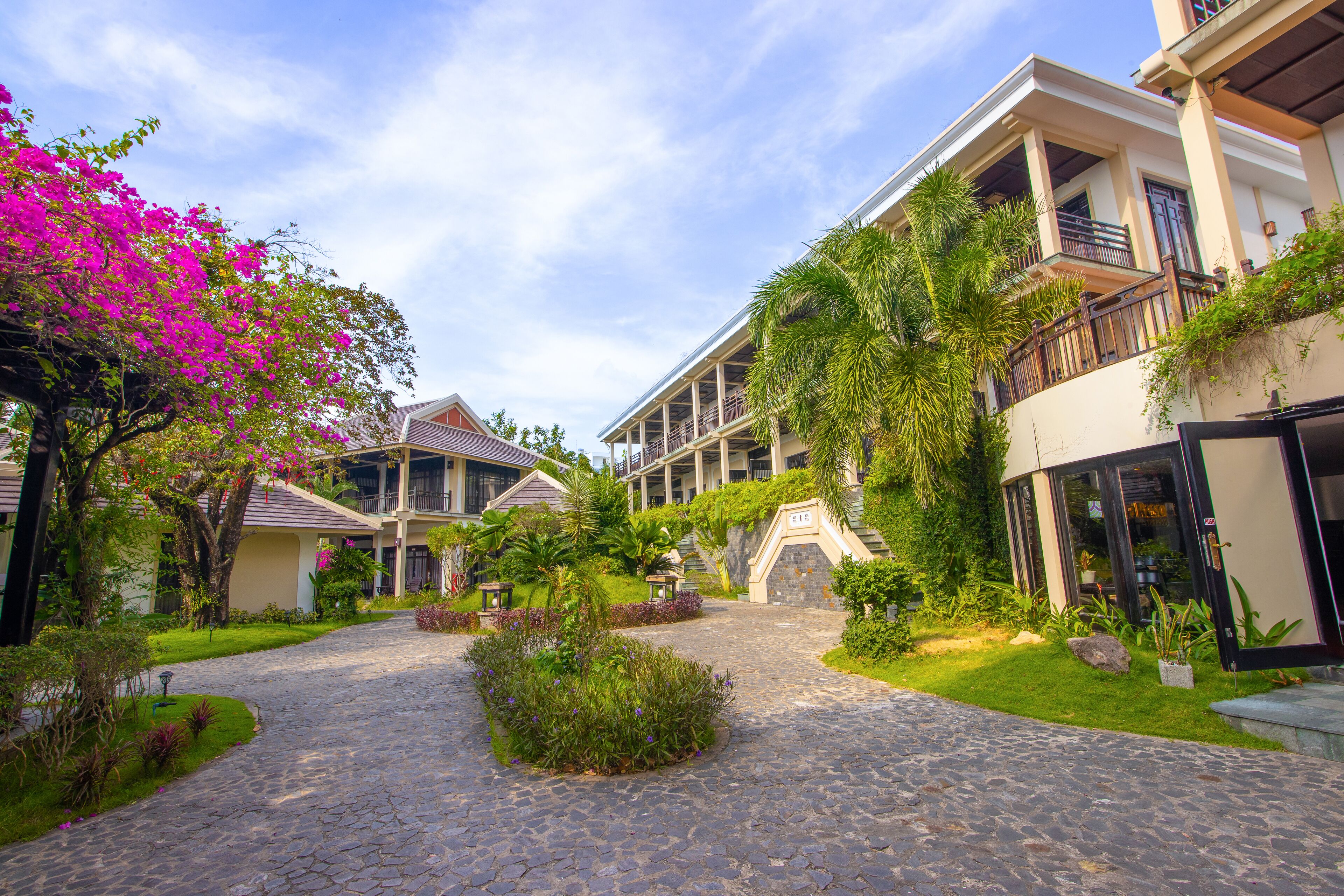 A courtyard with a cobblestone path, lush greenery, and a building with a balcony.