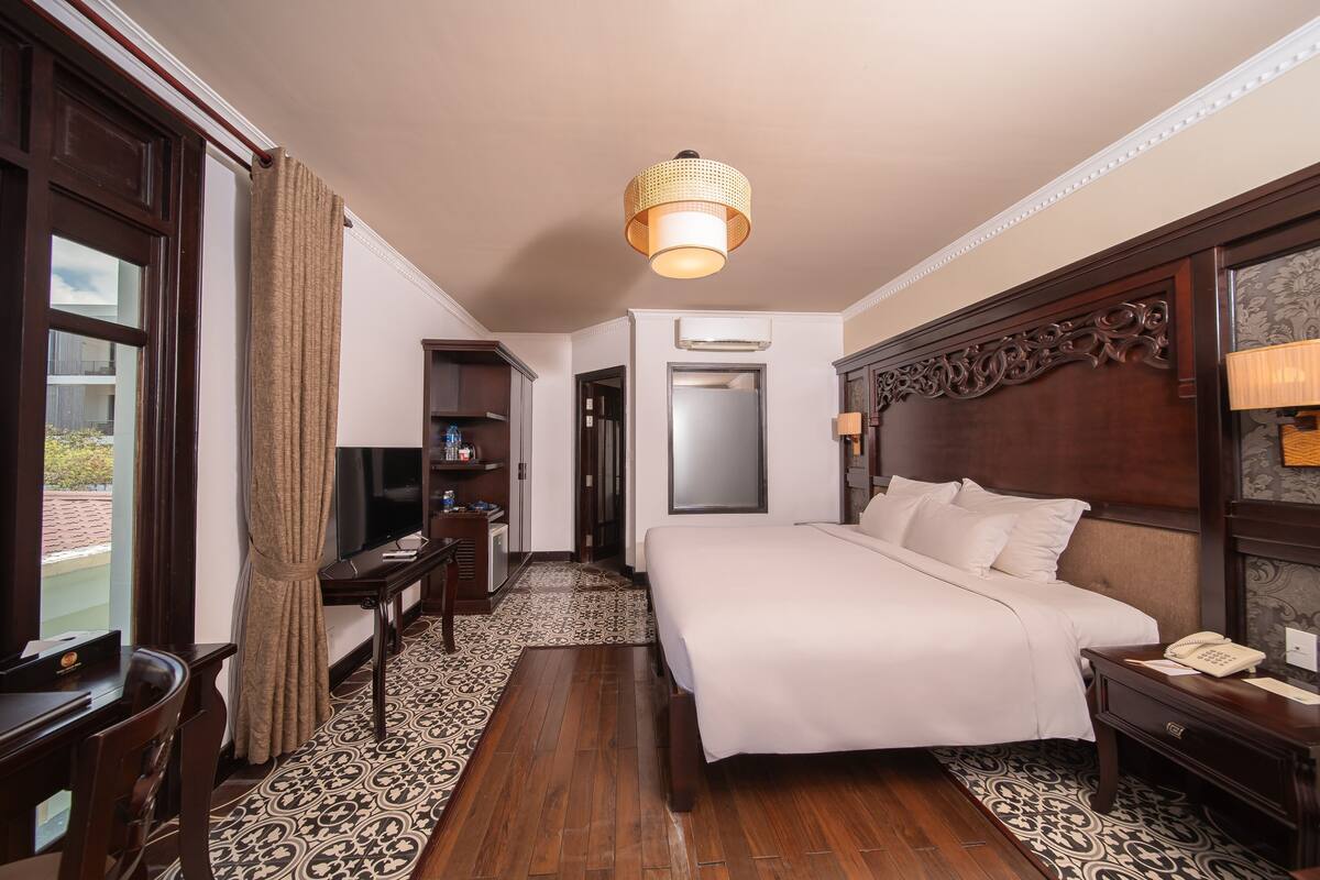 Camera d'albergo con un letto grande, una testiera in legno, una scrivania con una sedia, una televisione e un pavimento decorato.