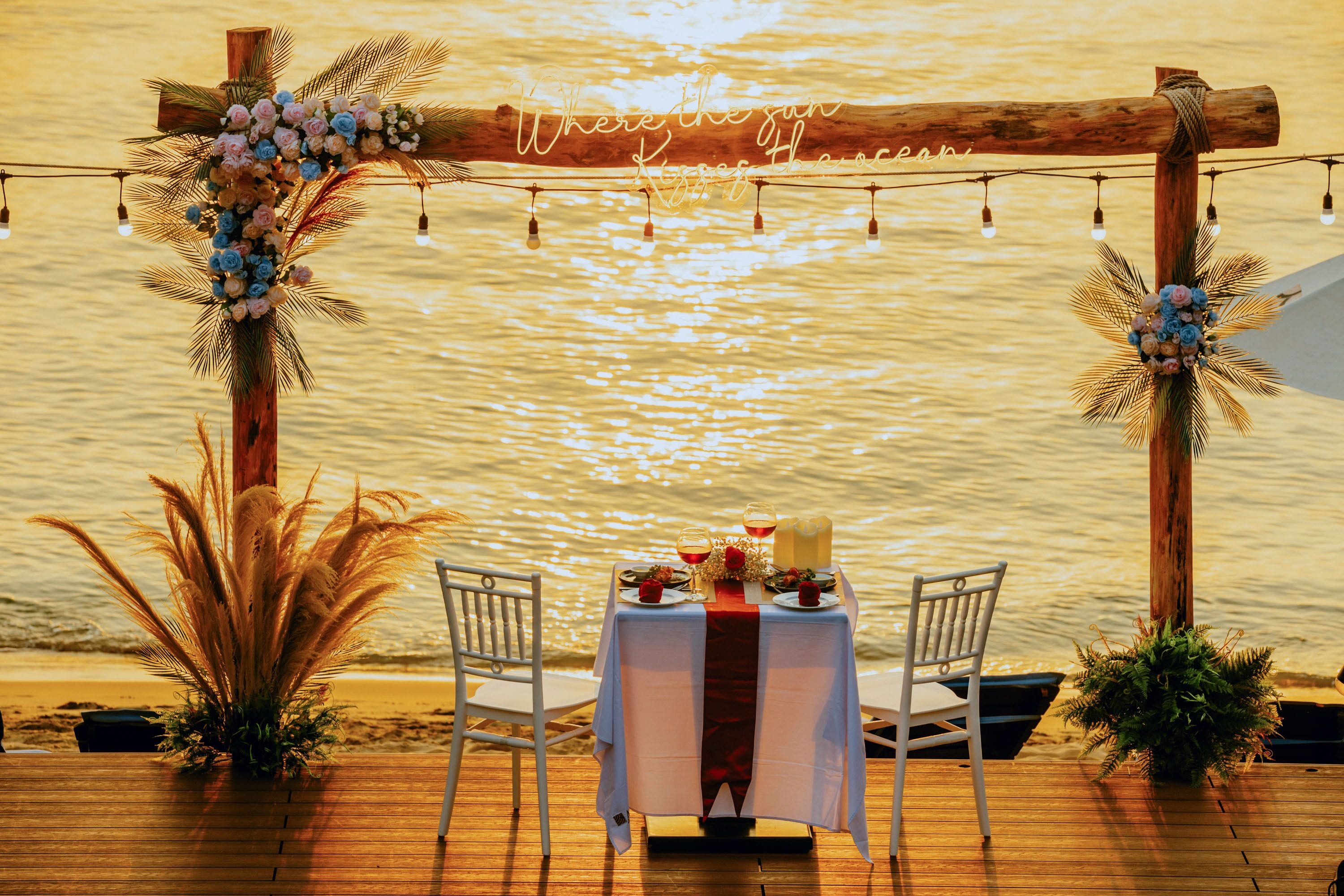Una terraza de madera con una mesa para dos, decorada con flores y un camino rojo, con vistas a una masa de agua.