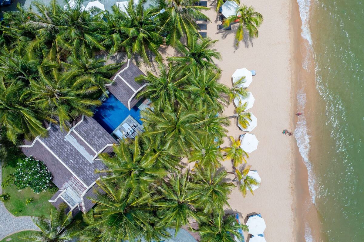 Una vista aerea di un resort sulla spiaggia, con palme, una piscina e ombrelloni.