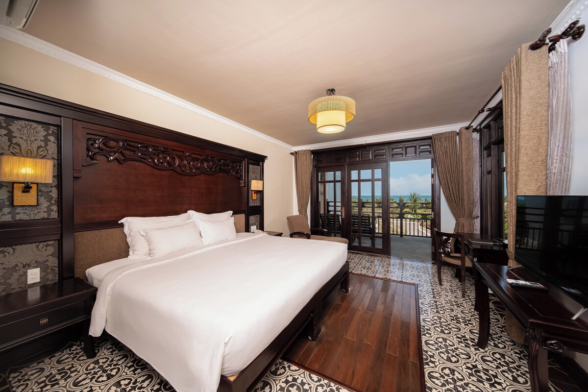 A hotel room with a large bed, a TV, a balcony with curtains, and a patterned floor.