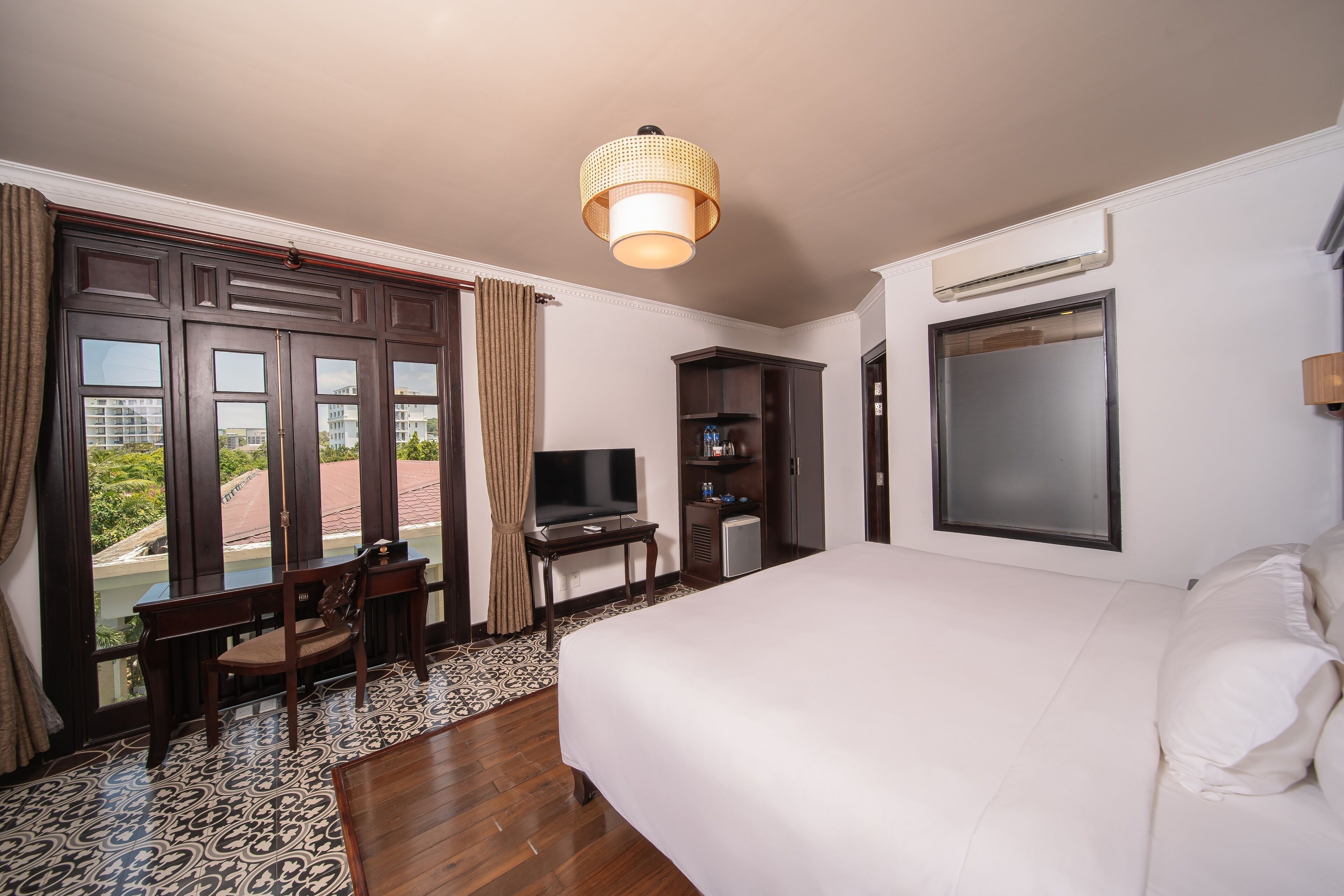Habitación de hotel con una cama grande, un escritorio con televisor, un refrigerador pequeño y una ventana con cortinas.