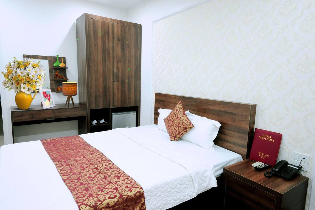 A hotel room with a bed, a nightstand with a phone and a directory, a desk with a vase of flowers, and a wardrobe.