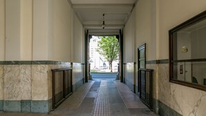 Un couloir aux murs de marbre, doté d’un sol carrelé, et offrant une vue sur une rue bordée d’immeubles et d’un arbre.