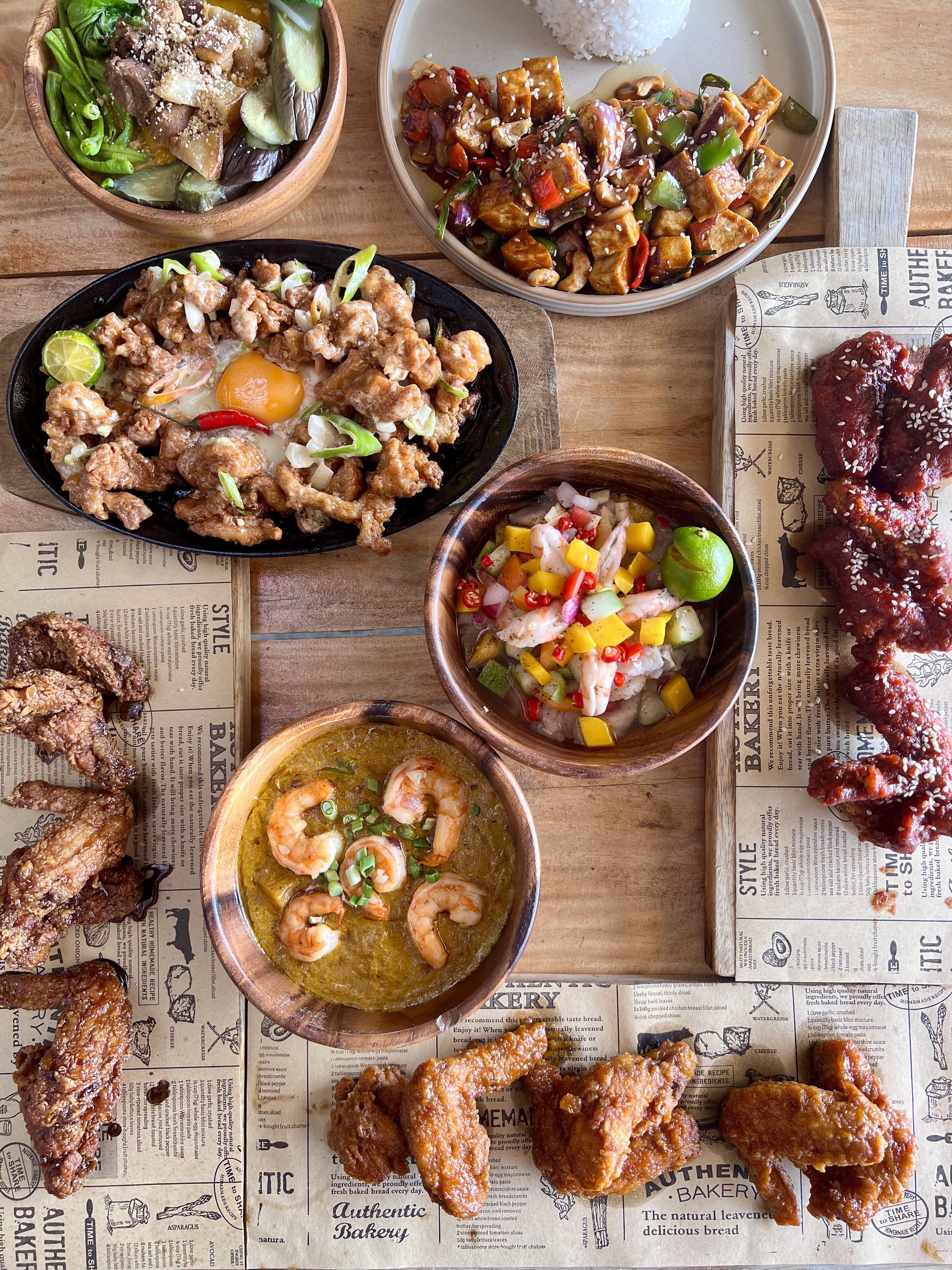 A variety of dishes including fried chicken, shrimp, and stir-fried vegetables, arranged on a wooden table with newspaper-style placemats.