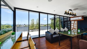A modern living room with a glass table, wooden flooring, a black sofa, and a view of the cityscape. - Casa Bella Phuket (Chalong)