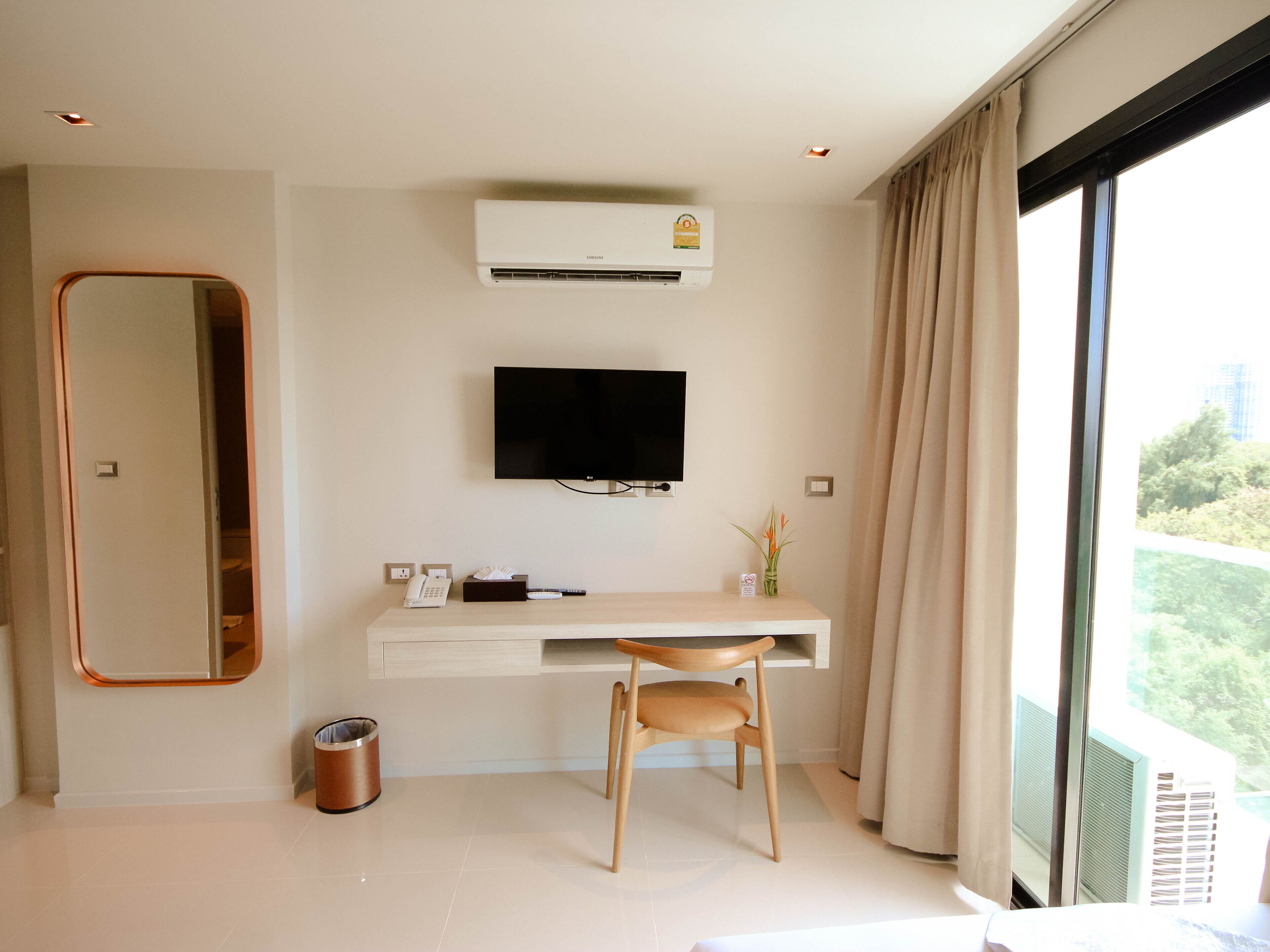 A modern hotel room with a large mirror, a flat-screen TV mounted on the wall, a wooden desk with a chair, a small trash bin, and a window with curtains.
