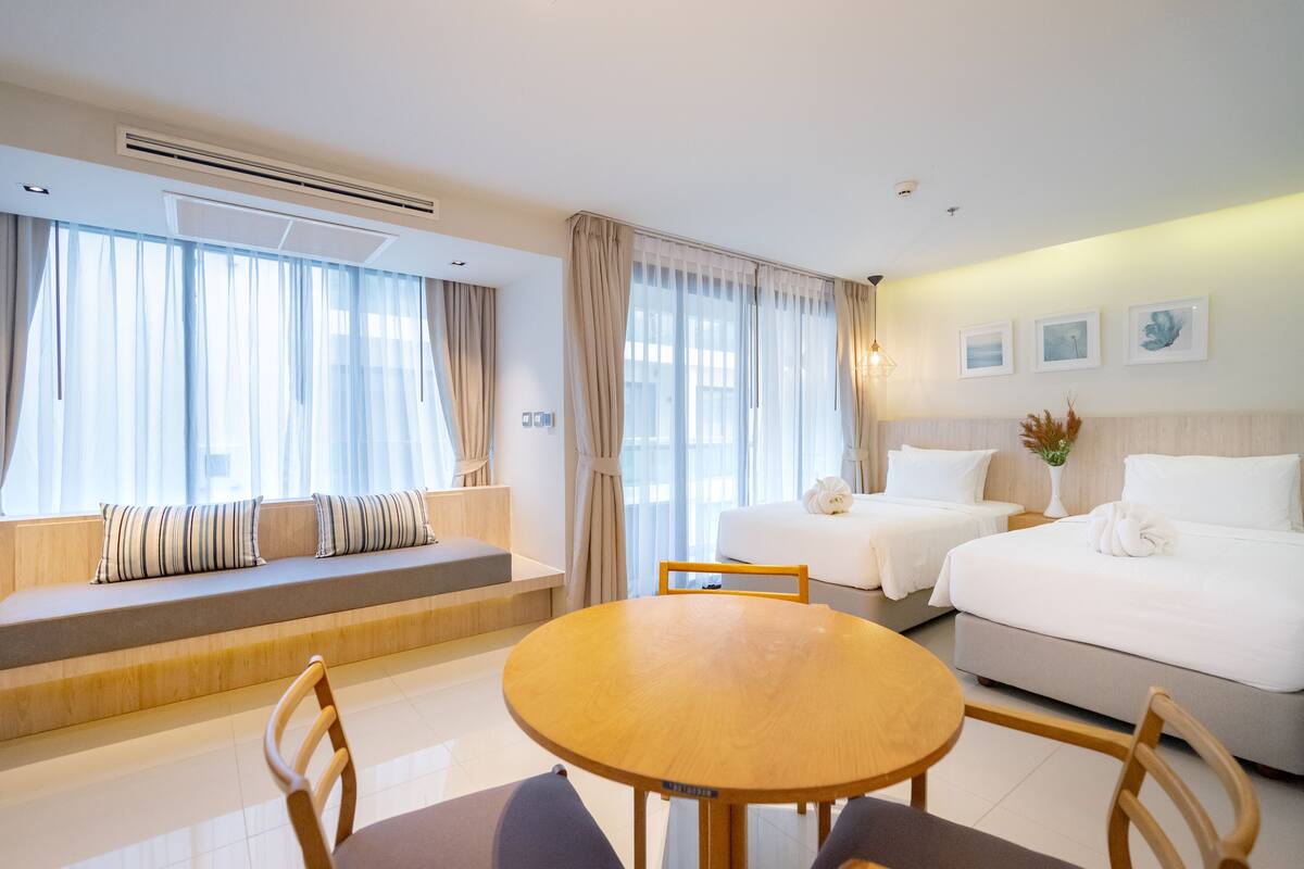 A hotel room with a large bed, a sofa bed, a dining table with chairs, and a window with sheer curtains.