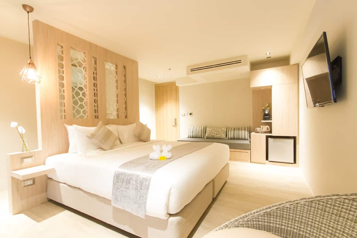 A modern hotel room with a large bed, a bedside table, a TV mounted on the wall, and a small kitchenette area.