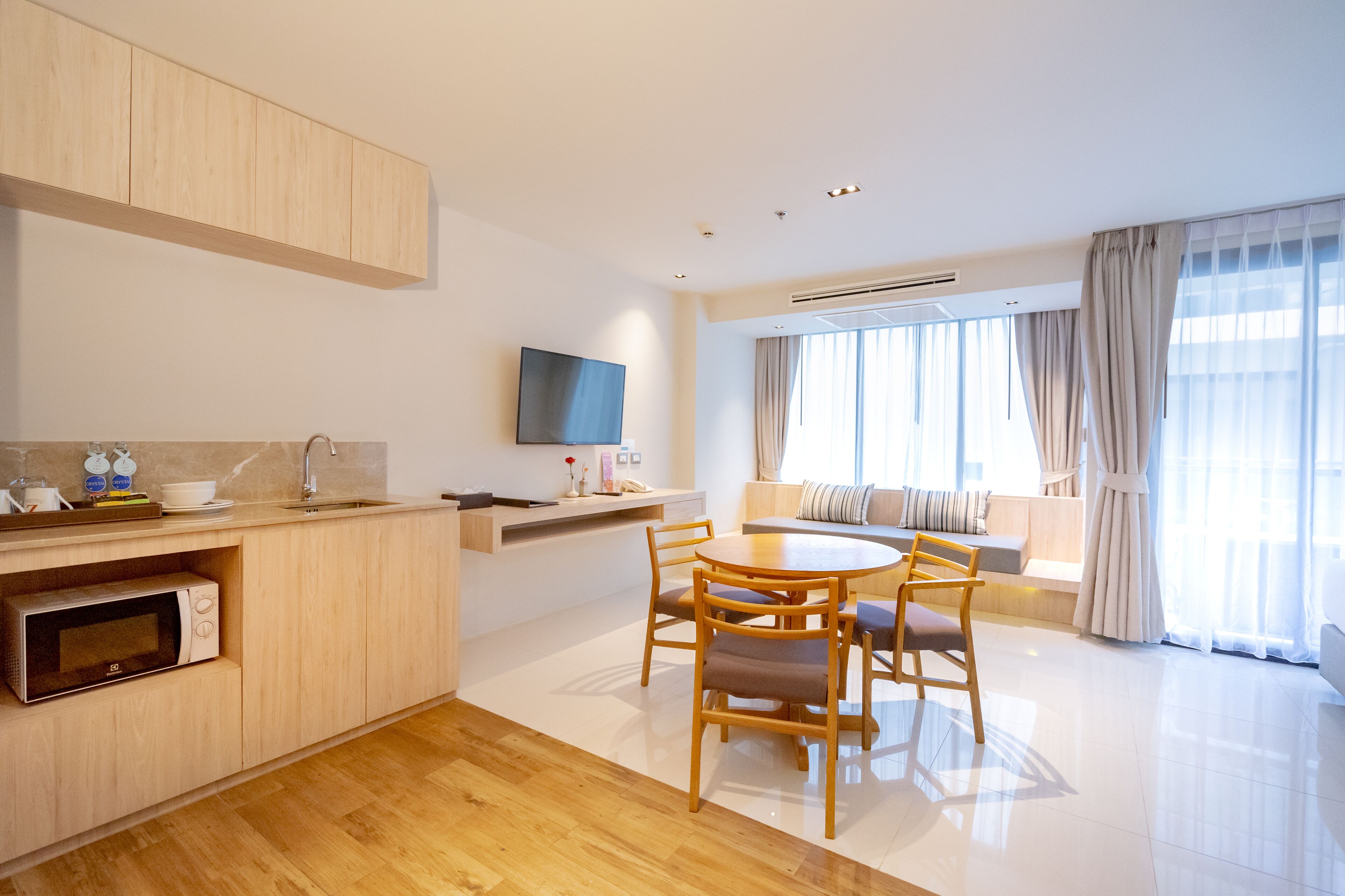 A modern kitchen with a microwave, a dining area with a round table and chairs, a flat-screen TV mounted on the wall, and a large window with curtains.