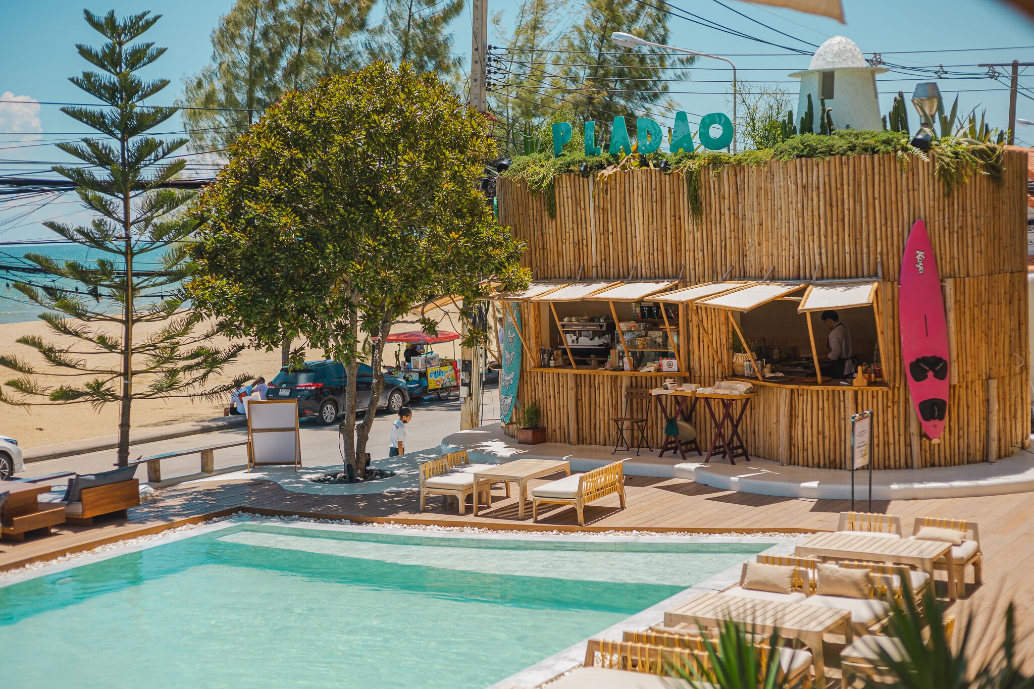 A poolside area with wooden lounge chairs, a bar with a surfboard decoration, and a view of the beach.