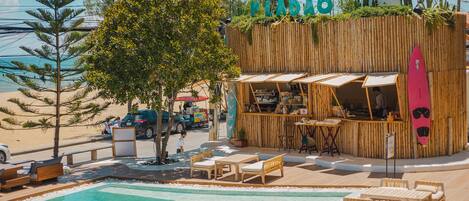 A poolside area with wooden lounge chairs, a bar with a surfboard decoration, and a view of the beach.