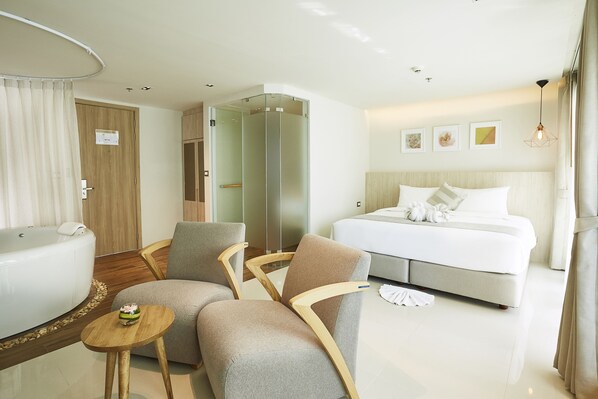A hotel room with a bed, two armchairs, a small table, a glass-enclosed shower, and a bathtub.