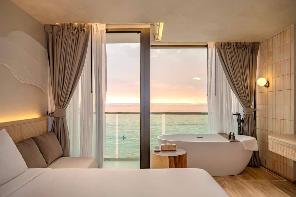 A modern hotel room with a large bed, a bathtub, and a balcony with a view of the sea.