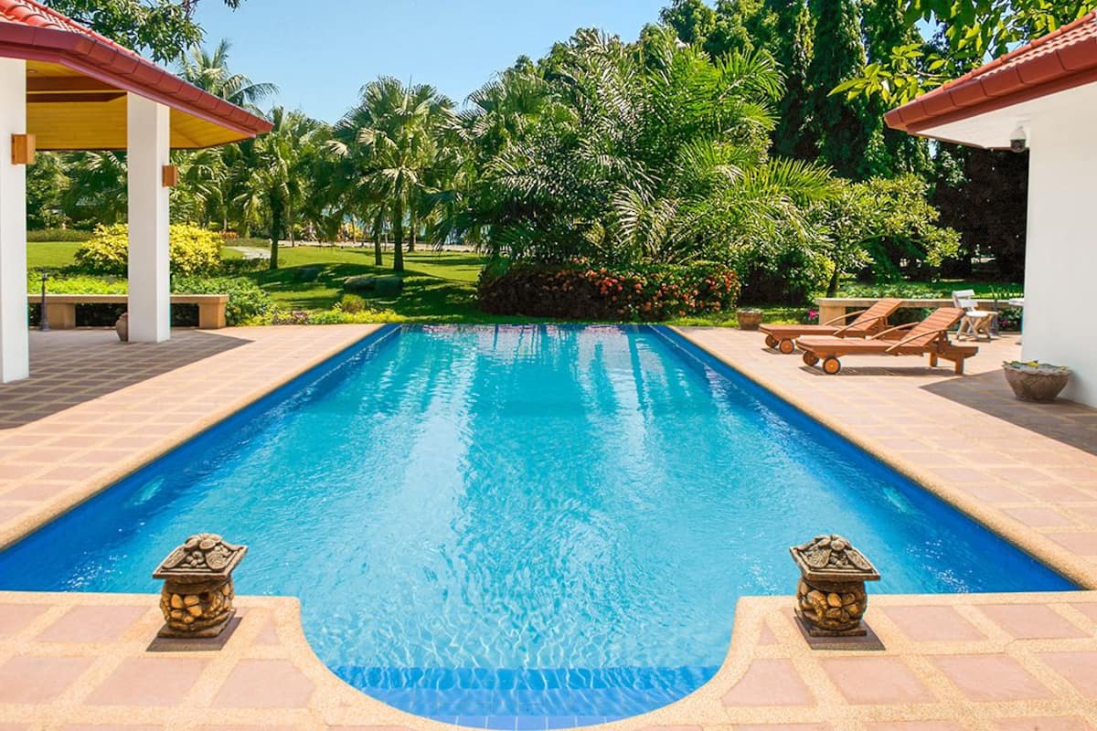 Piscina rectangular con agua azul cristalina, rodeada por un solárium con baldosas. Dos esculturas de piedra se encuentran a cada extremo de la piscina. La piscina está ubicada en un exuberante jardín con diversas especies de árboles y plantas.