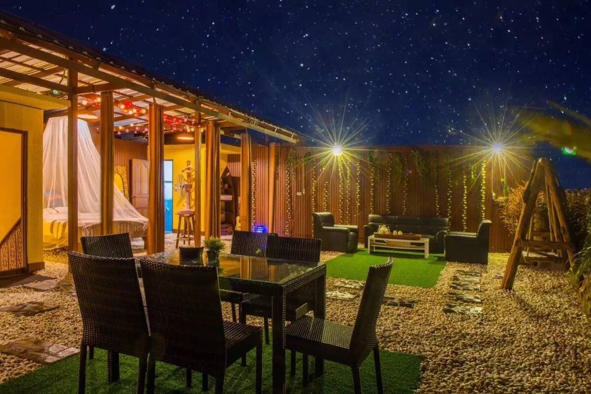 Un patio al aire libre con una mesa y sillas para comer, rodeado por una terraza de madera y luces colgantes.