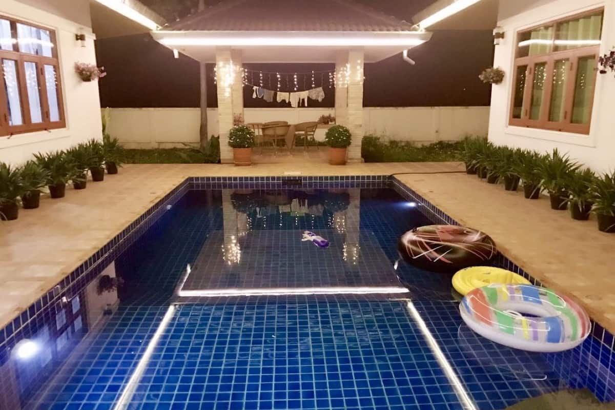 Una piscina con borde de baldosas, rodeada de plantas en macetas, y una zona cubierta con guirnaldas de luces.