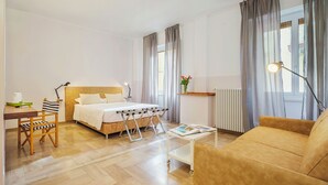 Una habitación de hotel con cama, sofá, una mesita, una silla, un radiador y una ventana con cortinas.