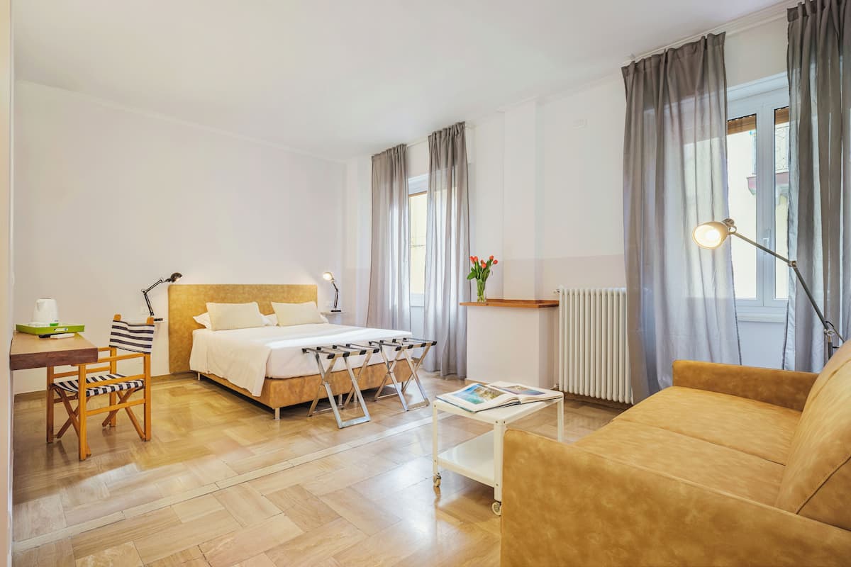 Una habitación de hotel con cama, sofá, una mesita, una silla, un radiador y una ventana con cortinas.