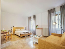 Una habitación de hotel con cama, sofá, una mesita, una silla, un radiador y una ventana con cortinas.