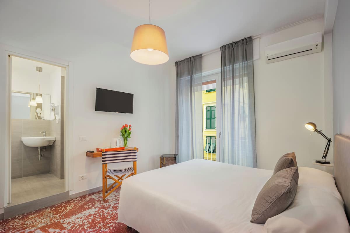 Habitación de hotel con cama, escritorio con silla, televisor montado en la pared, ventana con cortinas y baño con lavamanos y espejo.