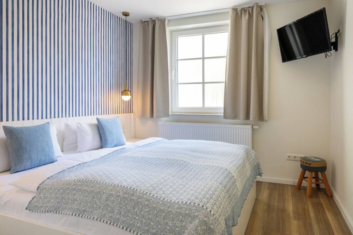 A bedroom with a bed, a window with curtains, a mounted TV, and a small stool.