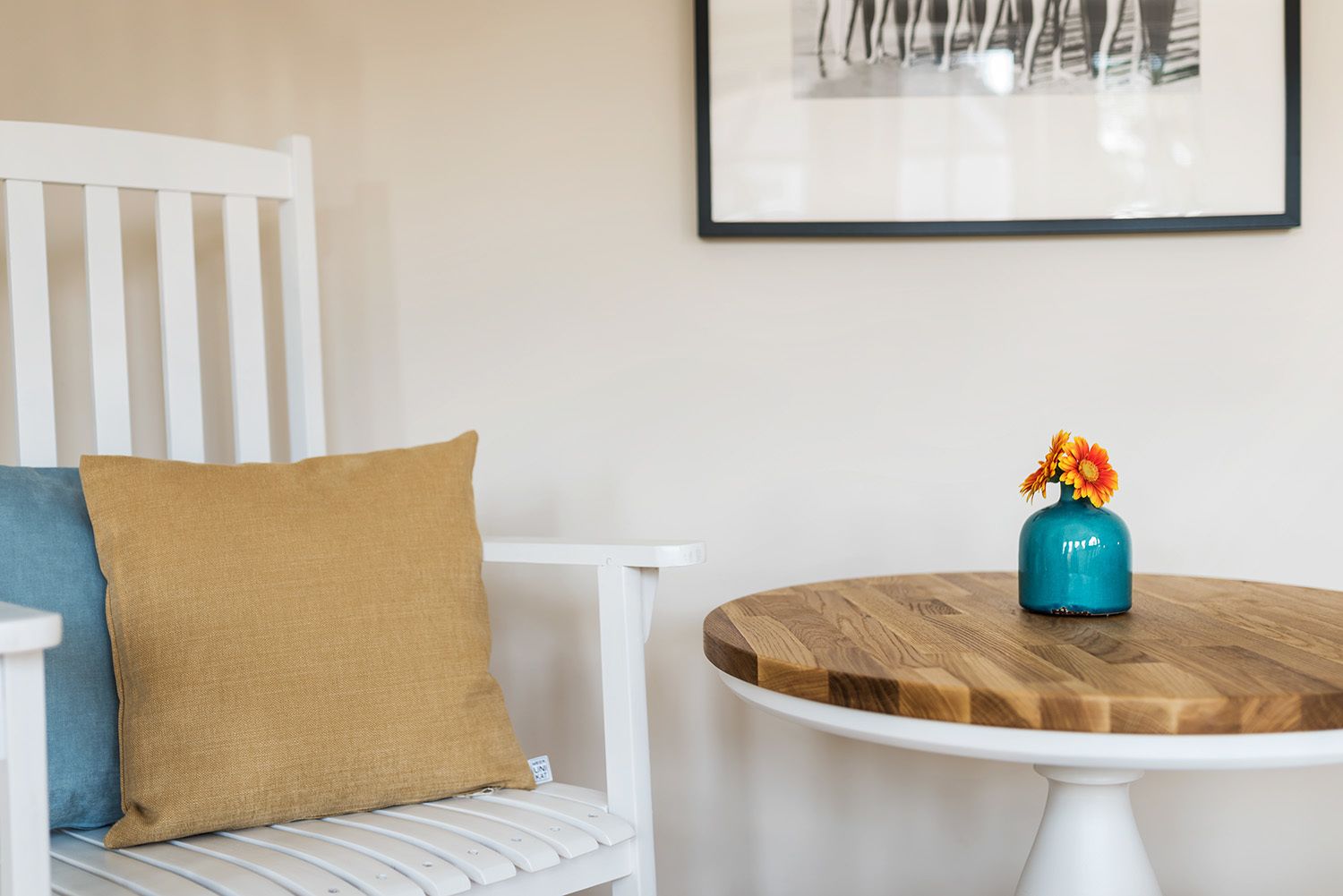 A white chair with a blue and mustard pillow, a small round wooden table with a blue vase and flowers, and a framed picture on the wall.
